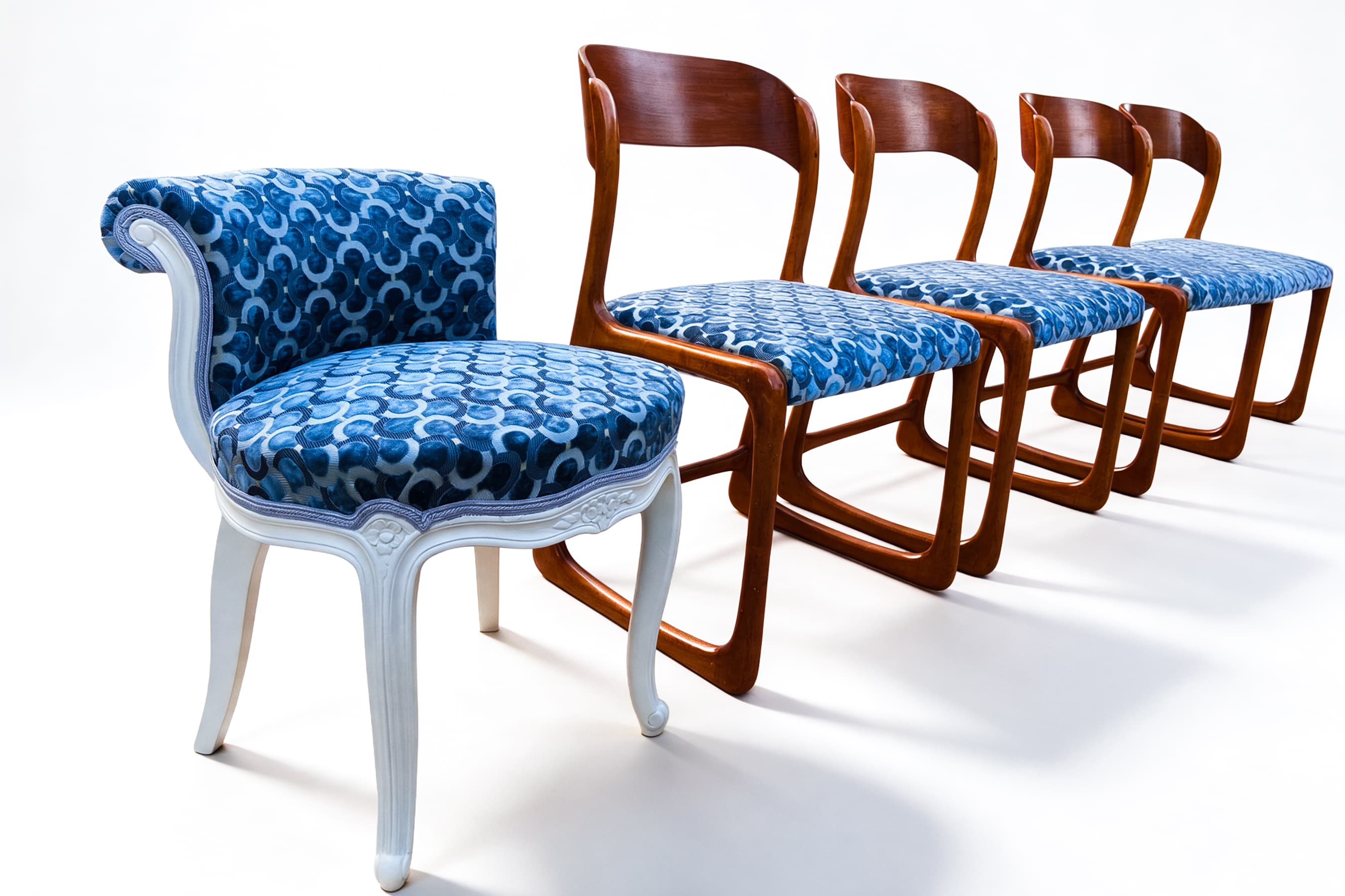 Une petite chaise laquée blanc au dossier enroulé et quatre chaises traîneau en bois, garnies d'un même tissu bleu à motif de cercles, sur fond blanc.