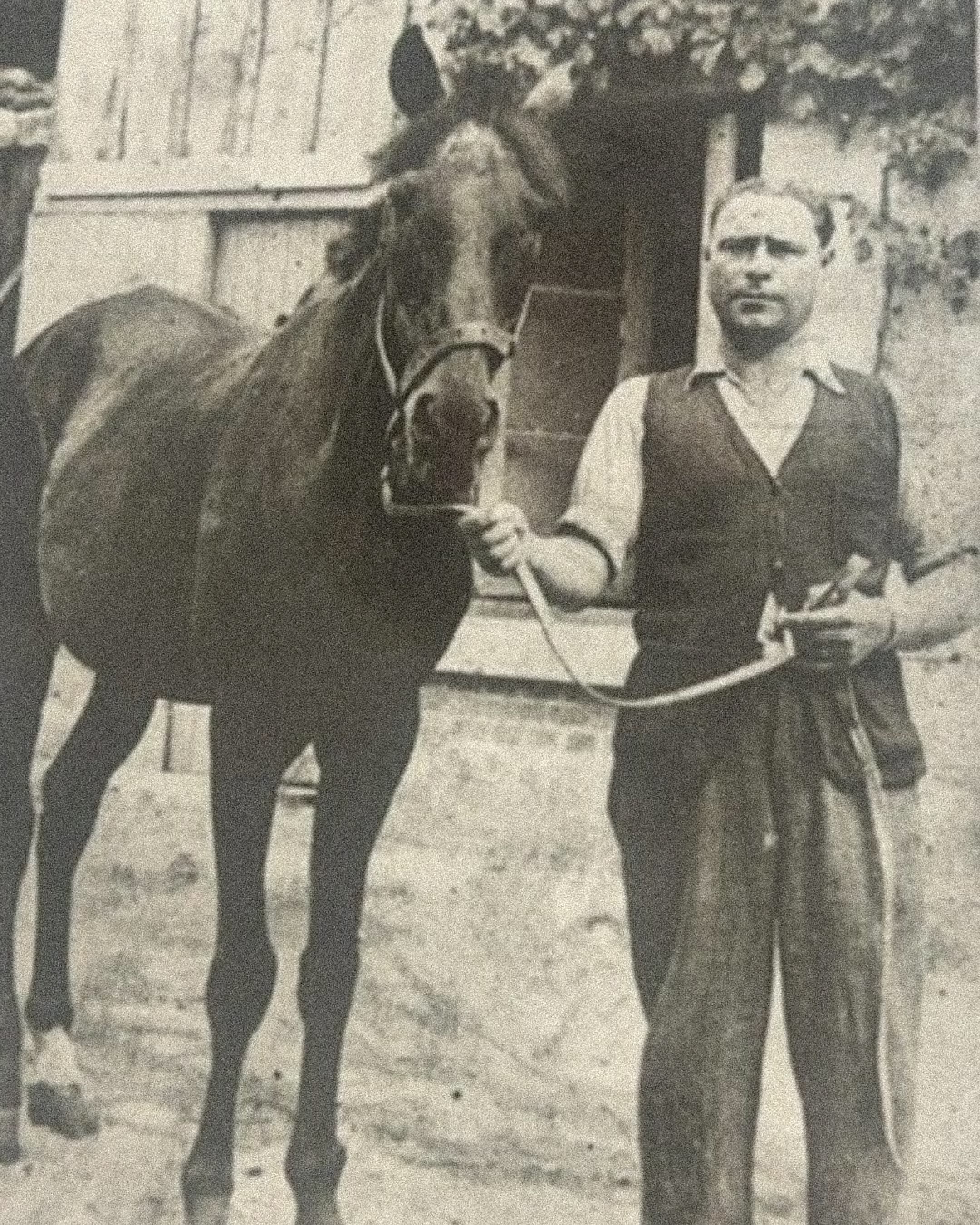 Photographie d'archive en noir et blanc : un homme en gilet et chemise aux manches retroussées tient un cheval par la longe, devant une bâtisse.