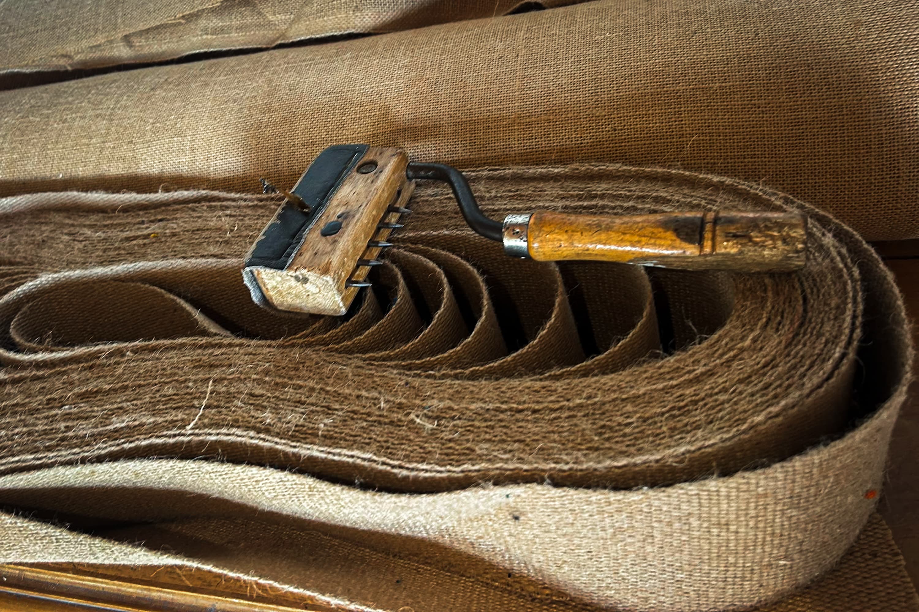 Un tire-sangle en bois posé sur un rouleau de sangle de jute, sur fond de toile.